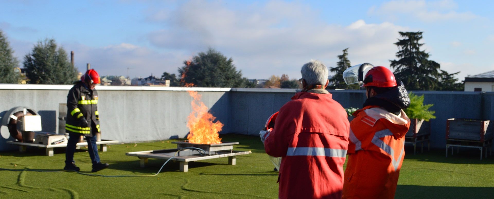 Addestramento antincendio sul tetto, con persone in tuta protettiva che osservano le fiamme.