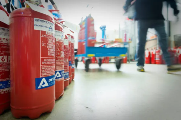 Estintori rossi allineati su un pavimento industriale, con una persona che cammina in secondo piano.