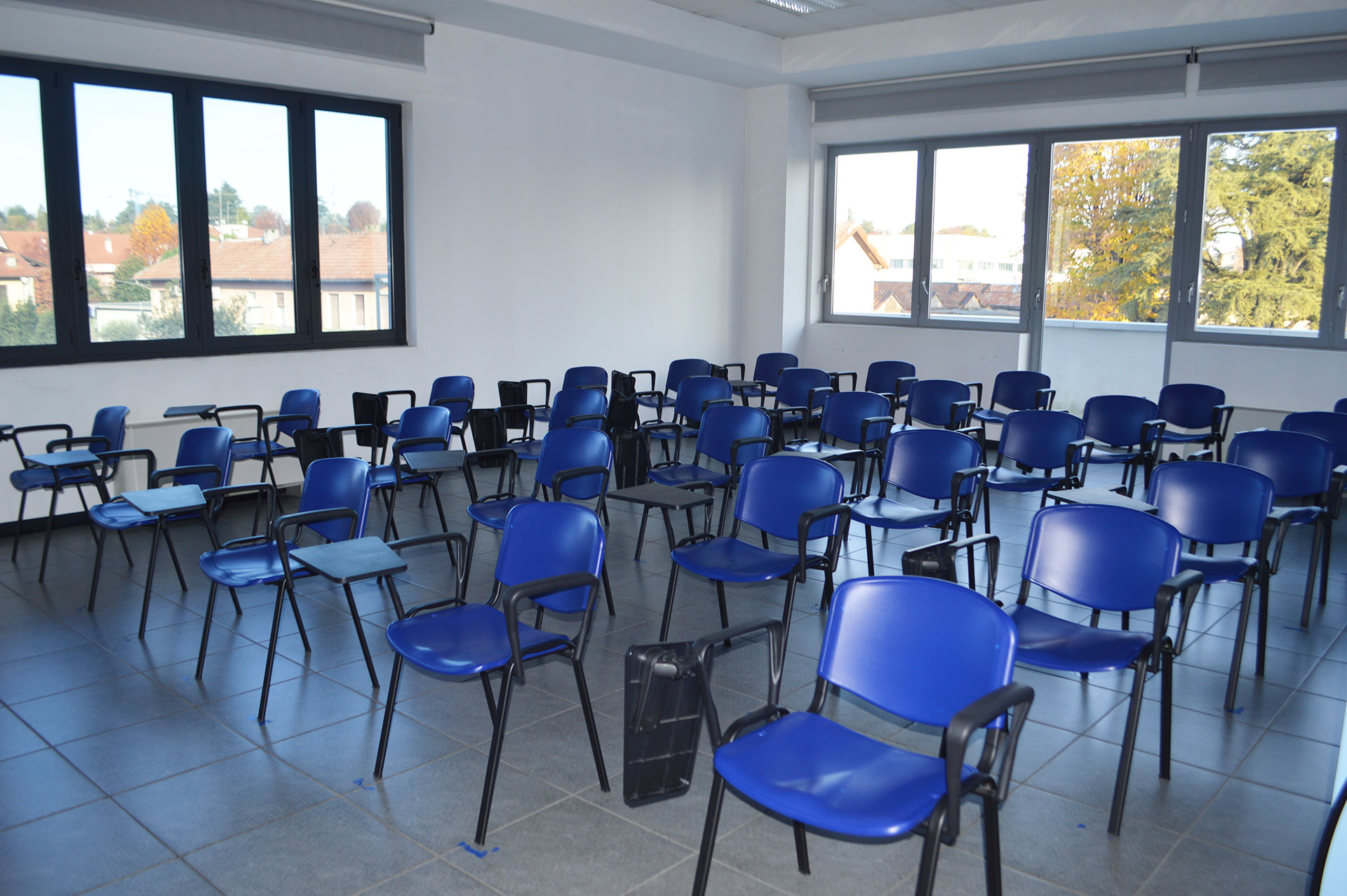 Aula vuota con sedie blu disposte in fila, ampie finestre che illuminano l'ambiente interno.