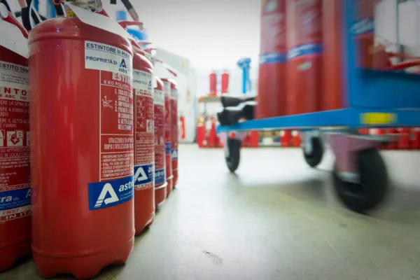 Estintori rossi disposti in fila e un carrello blu in movimento su un pavimento chiaro in un ambiente industriale.