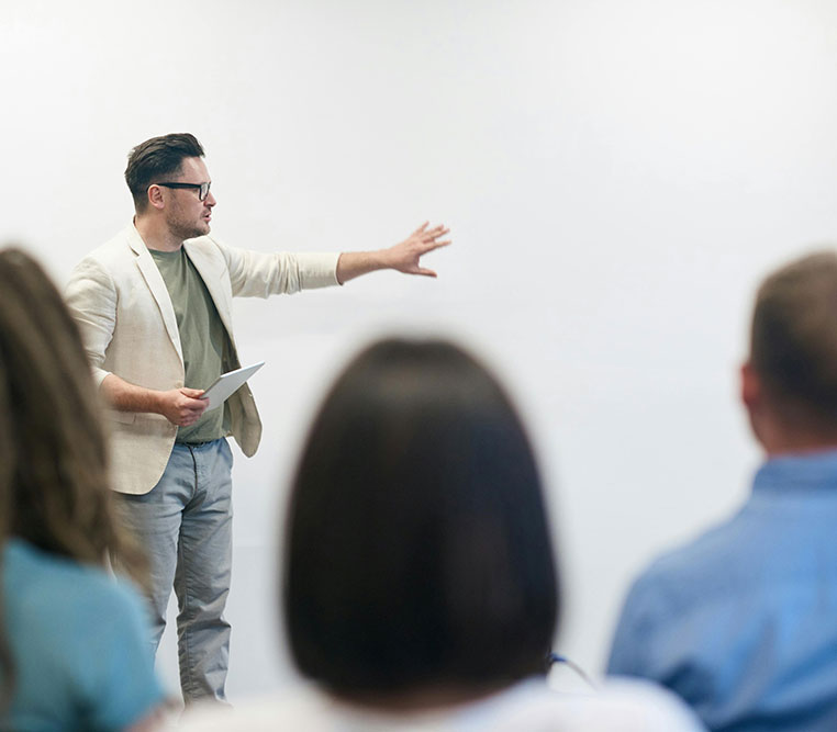 Uomo in abbigliamento casual tiene una presentazione davanti a un pubblico, spiegando concetti con un gesto del braccio.